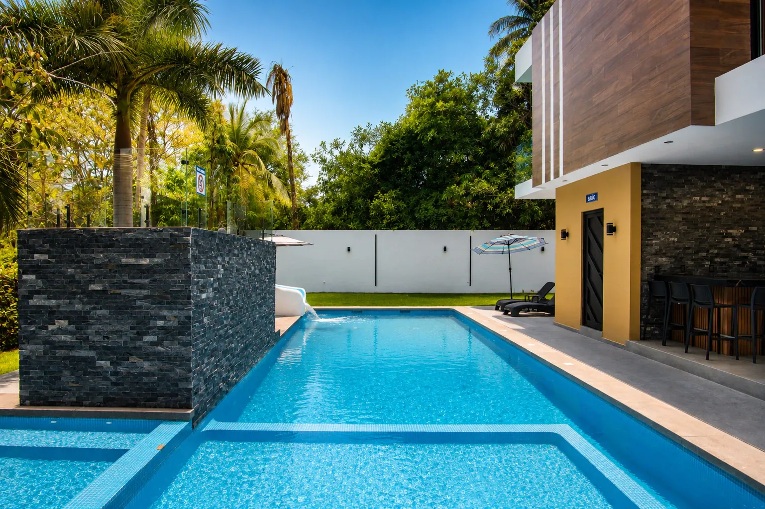 Alberca de mosaico azul junto a la palapa con barra y camastros, rodeada de palmeras